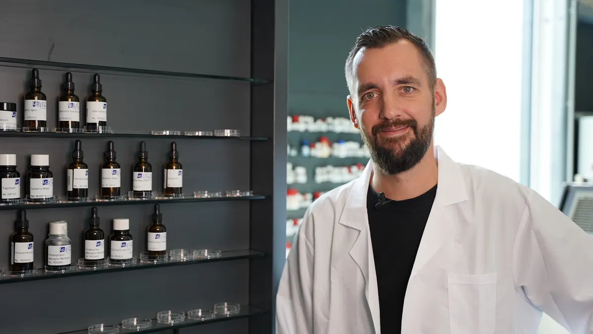 Perfumer in a white lab coat in front of a wall of shelves with fragrance bottles